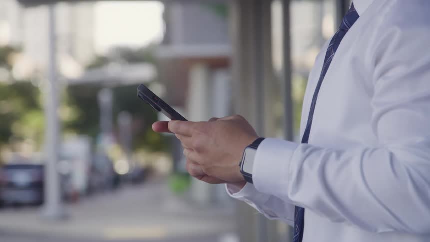 A closeup shot focuses on the torso and hands of a man in a white dress shirt and tie who is standing on a city street and actively typing on his black smartphone