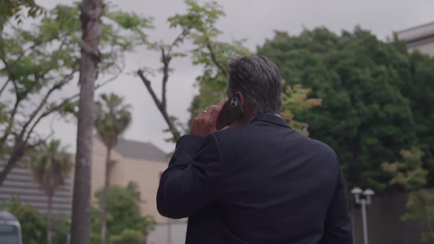 An adult male in a dark suit is looking forward while holding a phone to his ear standing outdoors surrounded by trees in a city on an overcast day