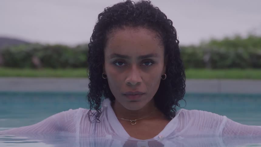 A young woman with black curly hair is in a swimming pool at midday the sunlight reflecting off the water around her