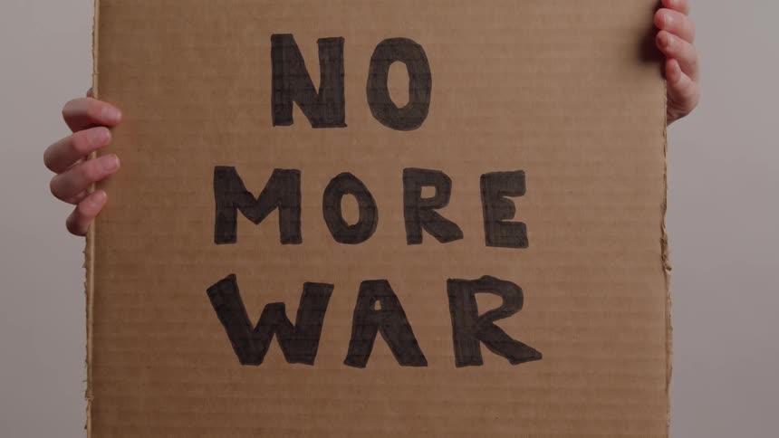 A pair of hands holds a cardboard sign reading no more war