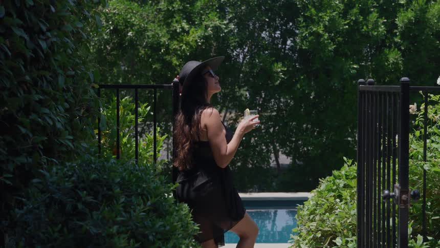 Young woman holds a drink as she stands in a park wearing a sunhat