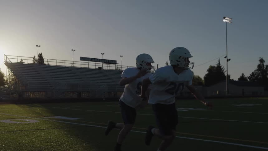 This clip shows two american football players wearing helmets and practice jerseys running a drill on an outdoor turf fieldthe players start in a crouched stance quickly run forward and one player catches a thrown football all silhouetted against
