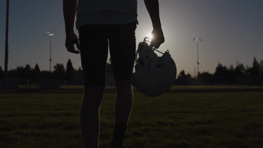 Football player walks across the field carrying his helmet in his hand