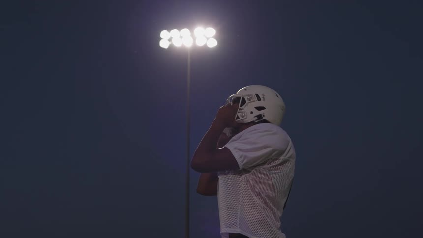 American football player adjusts his helmet on the field under floodlights at night