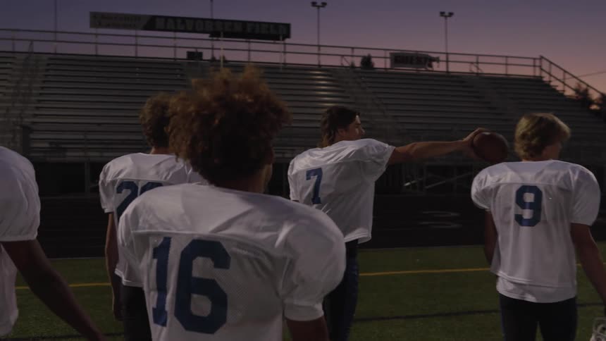 Several young male football players viewed from behind in white jerseys walk across a dark field towards empty stadium bleachers as the evening sky transitions from twilight to night