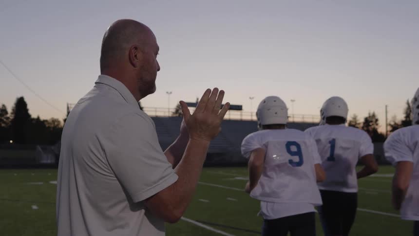Coach claps while american football players run across the field at dusk