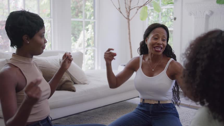 Three women sit on the floor of a bright living room smiling and snapping their fingers to a rhythm