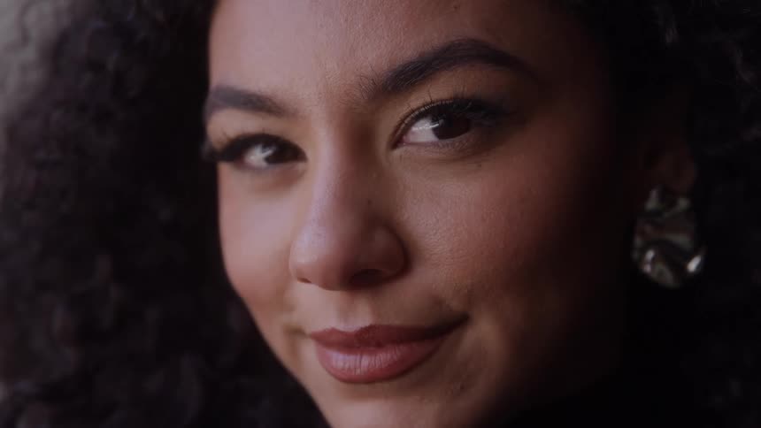 A closeup of a smiling woman looks directly into the camera against a dark background