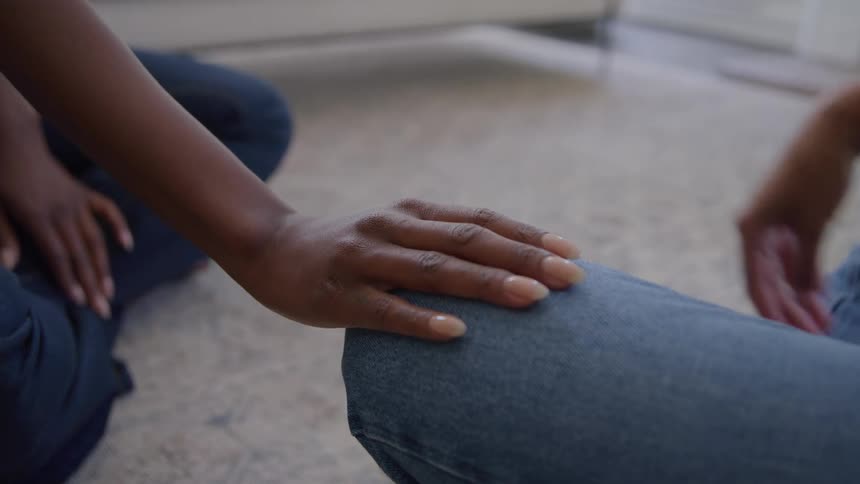 Person covers the hand placed by a second person on their knee as they sit on the living room floor