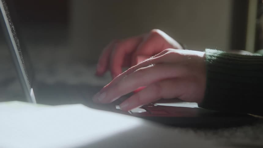 A close view of a person wearing a green sweater using the trackpad and keyboard of a laptop in a dimly lit space with a bright light source on the left