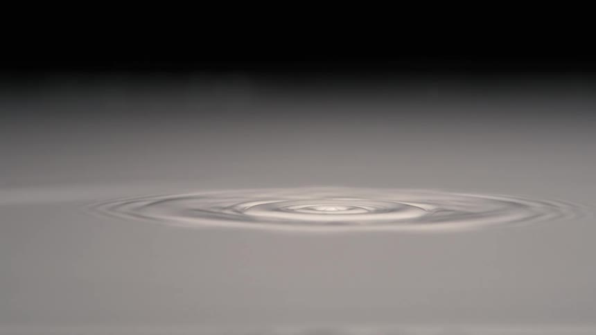The video captures a closeup slowmotion shot of a single water drop impacting a still pool of liquid the impact creates a classic coronet splash which then recoils to form a column that separates into another drop before the surface settles into expa