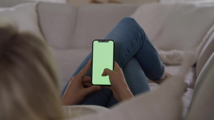 Woman sitting on the couch scrolling on her cell phone with a green screen