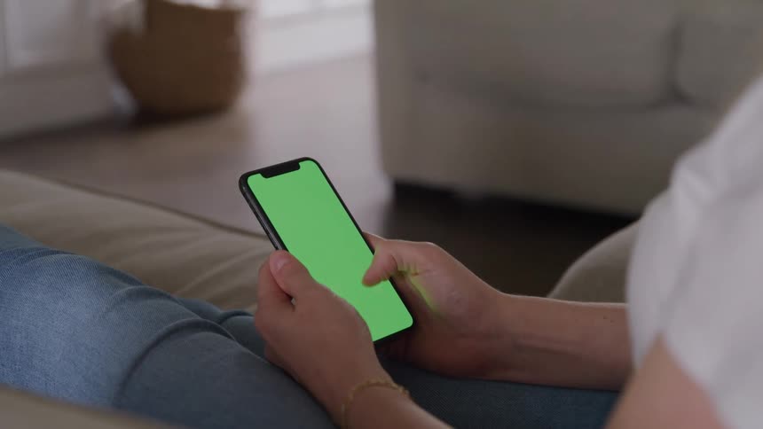 A person sits on a couch and scrolls on a smartphone with a green screen