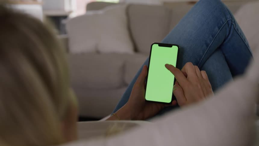 A young woman lies comfortably on a couch in her living room operating her mobile device with focus and ease