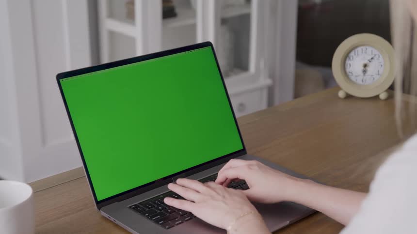 Woman sitting at a table typing on a laptop computer with a green screen