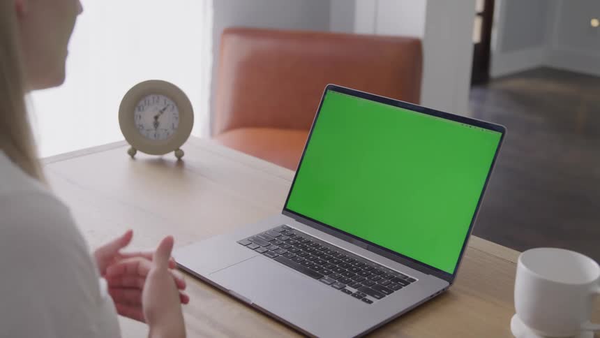 Woman sitting at a table in front of a laptop computer with a green screen as she hold her hands up and talks