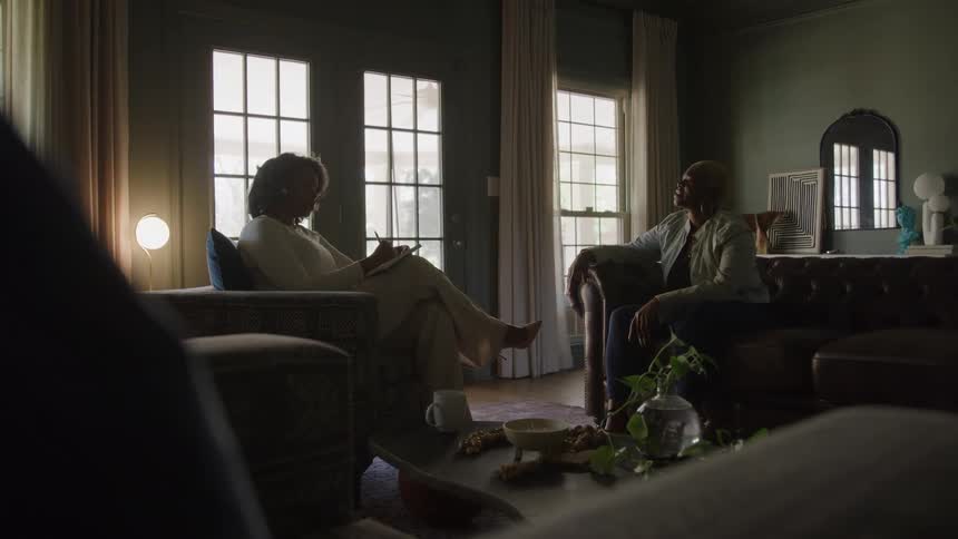 The scene features two women sitting across from each other in a dimly lit stylish living room with large french doors letting in natural light one woman is seated on a leather sofa while the other sits in a nearby armchair writing in a notepad sugge