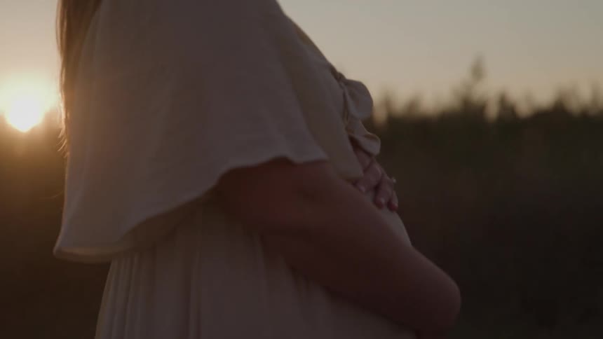 Pregnant woman holds her belly while standing in a field at sunset