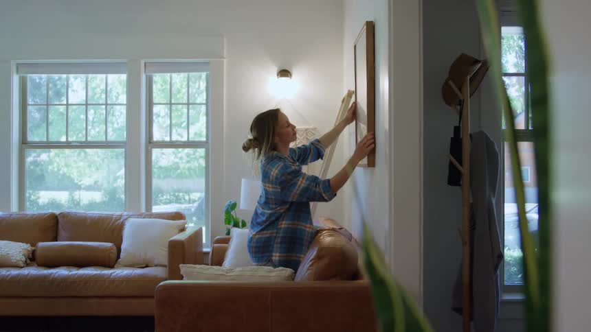 A woman wearing a blue plaid shirt stands on a leather couch in a bright living room to hang a framed picture on a white wall next to a sconce light