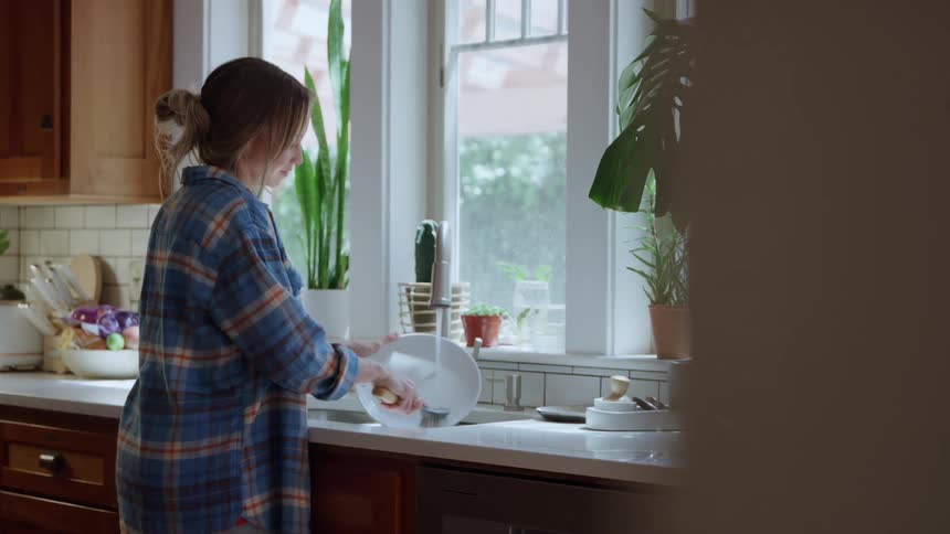 Woman washes white plate at kitchen sink lit by natural light