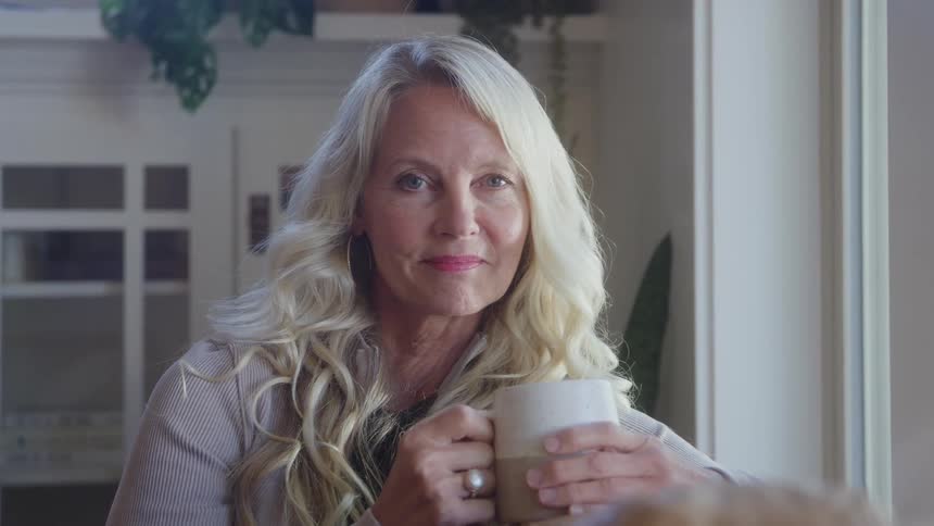 Adult woman with blonde hair sitting down and smiling while holding a cup with her hands indoors during day time