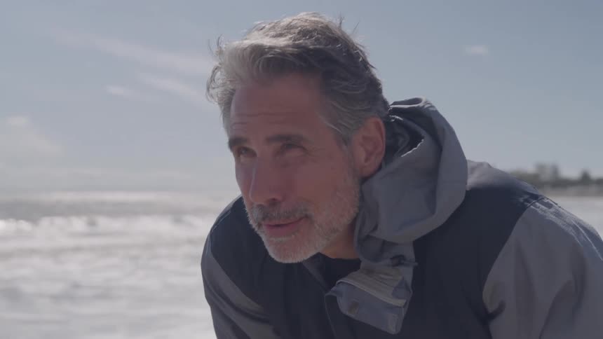 Man with beard looks at coastline under clear sky wearing gray and blue jacket