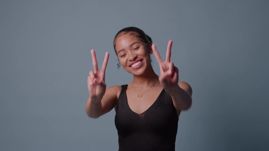 Woman smiles making peace sign with both hands against bluegray background