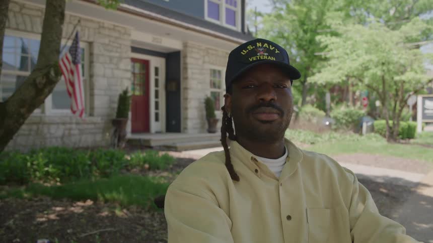 Man wearing a us navy cap positioned in front of a house