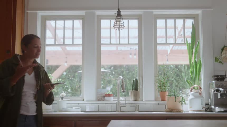 Woman walks back and forth across the kitchen holding out her phone and talking as she moves her arm up and down