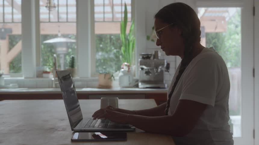 Woman with braid works on laptop at a bright kitchen counter