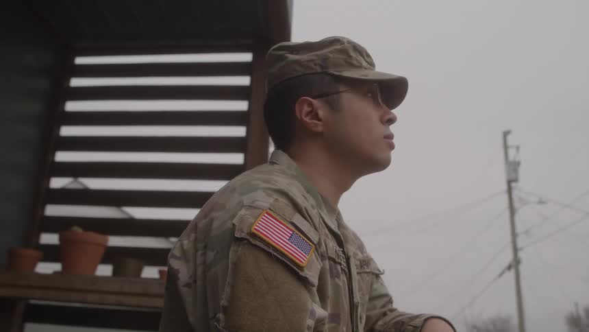 A young military male officer dressed in his uniform and wearing a hat sits on the front porch at midday he gazes thoughtfully at his surroundings