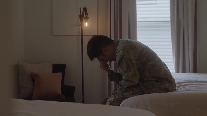 Service member sitting on the edge of the bed leans forward with hand over face