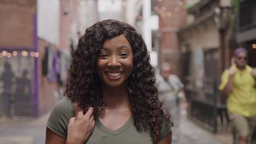 Woman standing outdoors between urban buildings smiles as pedestrians walk by