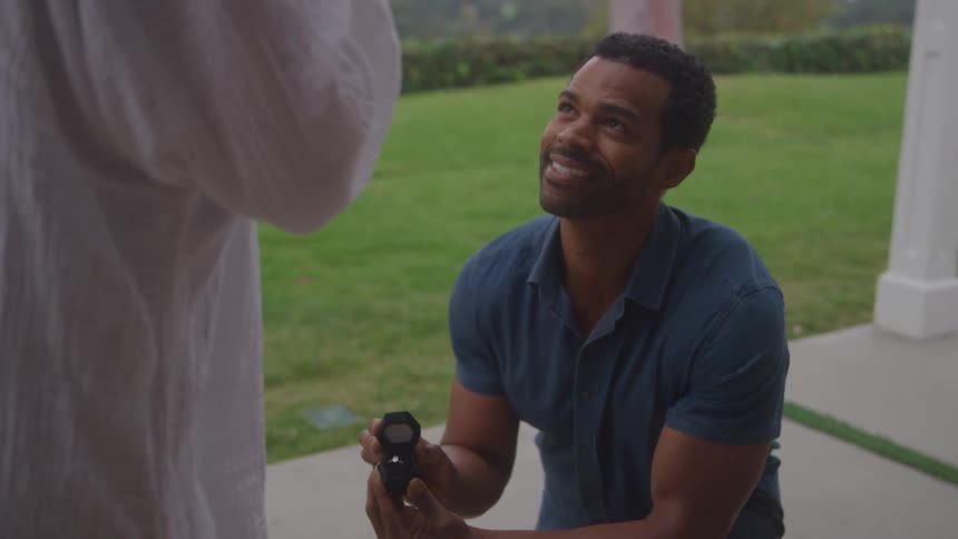 A smiling man kneels with an open ring box as he makes a marriage proposal outside a home