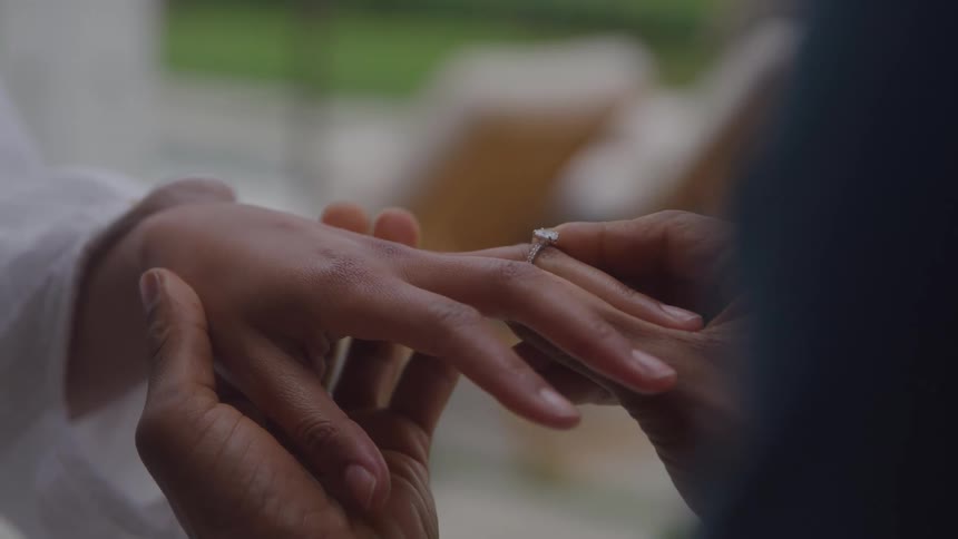 In a tender closeup a pair of hands gently slides a diamond ring onto a finger while the soft outoffocus background suggests a quiet outdoor setting