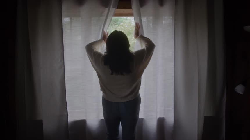 Woman opens sheer curtain at window with view of trees wearing light sweater and jeans