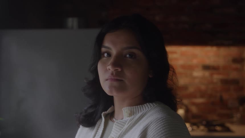 Woman with dark hair and white sweater stands indoors with brick wall and kitchen behind warmly lit