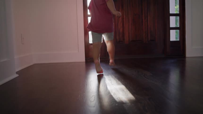 Child walking barefoot on dark wooden floor with sunlight casting shadows on the ground