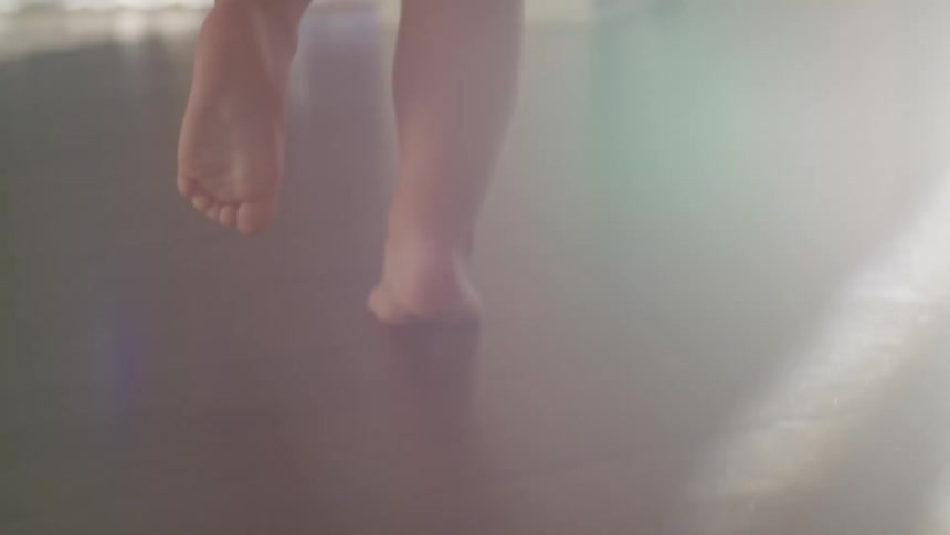 A low angle view of a young childs bare feet running across a dark hardwood floor moving past scattered toys and toward a bright sunlit doorway