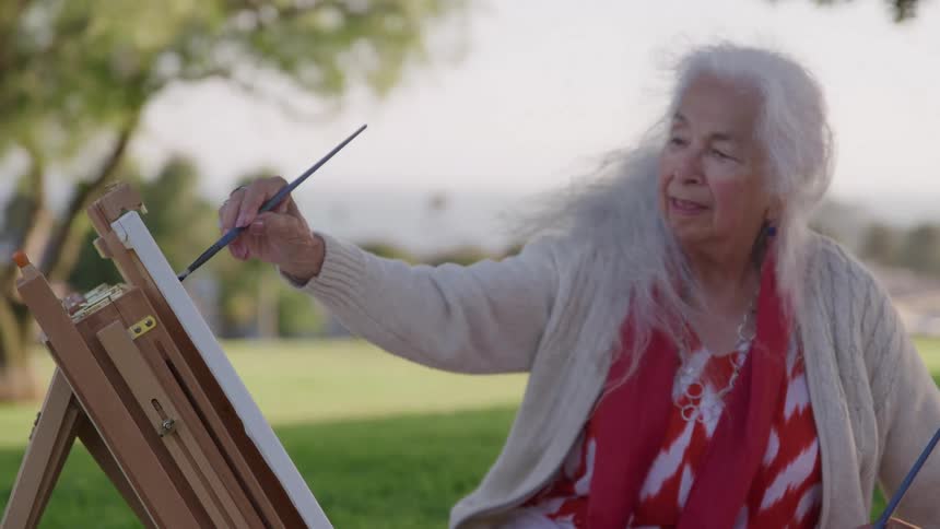 Elderly woman holding a paintbrush while painting on an outdoor easel
