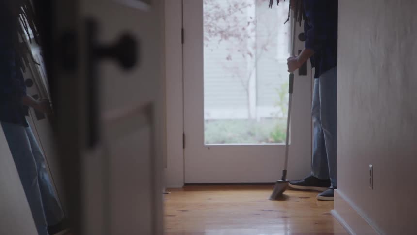 A man wearing jeans and a dark shirt sweeps a wooden floor near a glass paneled door that shows a blurred outdoor view