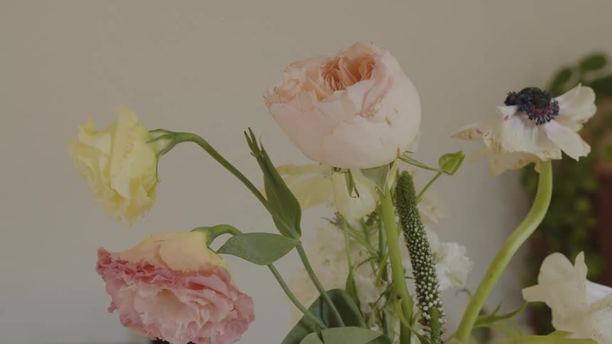 The arrangement features several prominent flowers in soft pastel colors including a large pale peachpink roselike bloom a ruffled pink flower and a slightly yellow bloom alongside smaller white flowers and green foliage