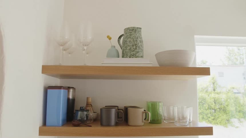 Floating wooden kitchen shelves hold various glasses mugs and decorative items with a bright sunny window in the background