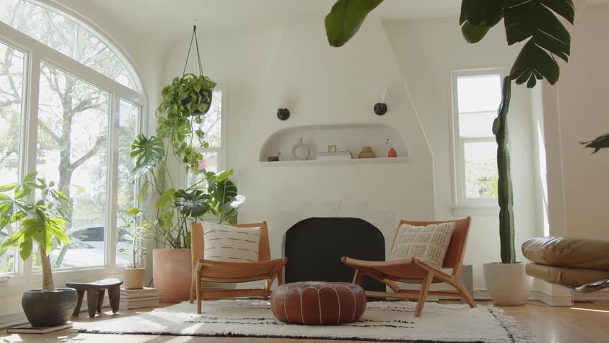 Bright natural light floods a white bohemian living room highlighting a kiva fireplace leather armchairs and an array of lush potted plants near a large arched window