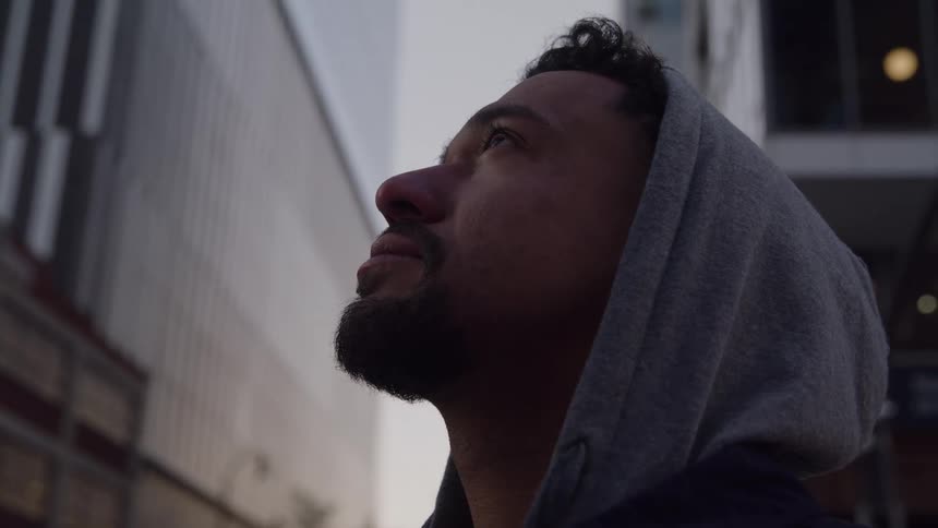 An adult male with a beard and a grey hood over his head is looking forward and upward against the backdrop of tall city buildings at dusk