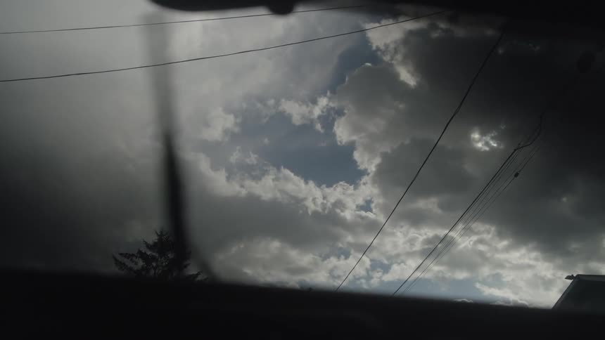 A windshield wiper sweeps across the glass while a cloudy sky partially veils the sunlight casting a soft diffused glow and faint reflections across windshield