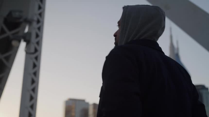 Hooded person walks beneath a metal structure with buildings in the background