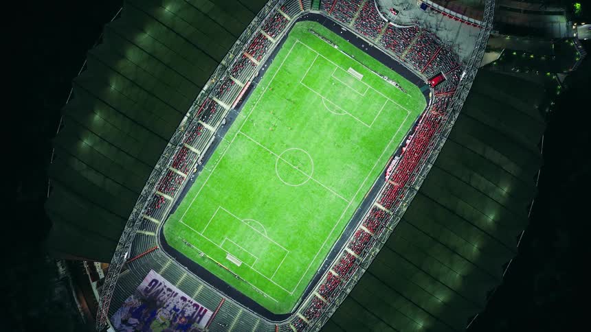 An aerial camera twists and rises high above a brightly lit soccer stadium at night transforming the intense game below into a distant glowing spectacle