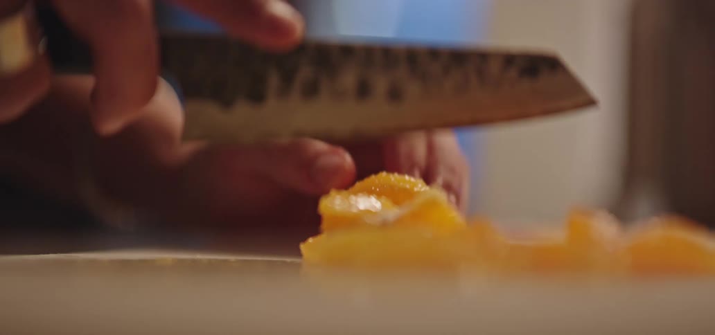 A closeup shot focuses on hands using a textured chefs knife to quickly slice bright orange food pieces into small cubes on a cutting board with the background remaining blurred