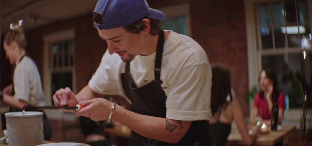 Waiter wearing a blue cap grating cheese over dishes of food with another waiter and clients at a table in the background indoors during night time